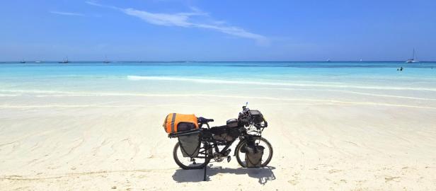 Mann mit Fahrrad und drei Kindern am weißen Sandstrand vor türkisblauem Meer und Segelbooten.