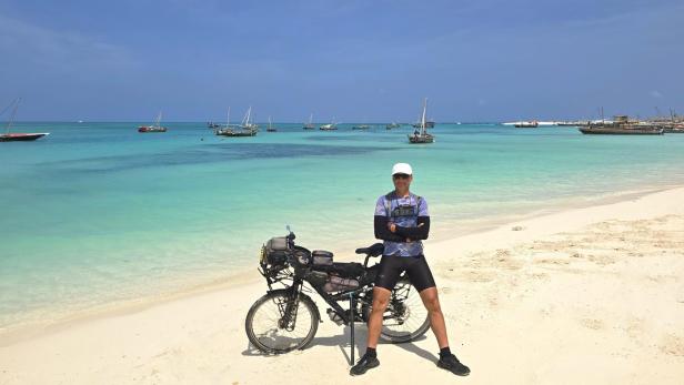 Mann mit Fahrrad und drei Kindern am weißen Sandstrand vor türkisblauem Meer und Segelbooten.