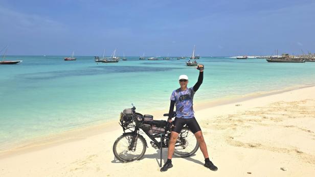 Mann mit Fahrrad und drei Kindern am weißen Sandstrand vor türkisblauem Meer und Segelbooten.