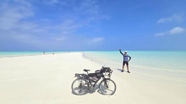 Mann mit Fahrrad und drei Kindern am weißen Sandstrand vor türkisblauem Meer und Segelbooten.