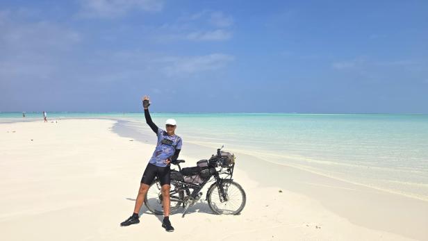 Mann mit Fahrrad und drei Kindern am weißen Sandstrand vor türkisblauem Meer und Segelbooten.