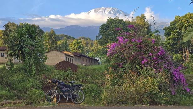 Mann mit Fahrrad und drei Kindern am weißen Sandstrand vor türkisblauem Meer und Segelbooten.