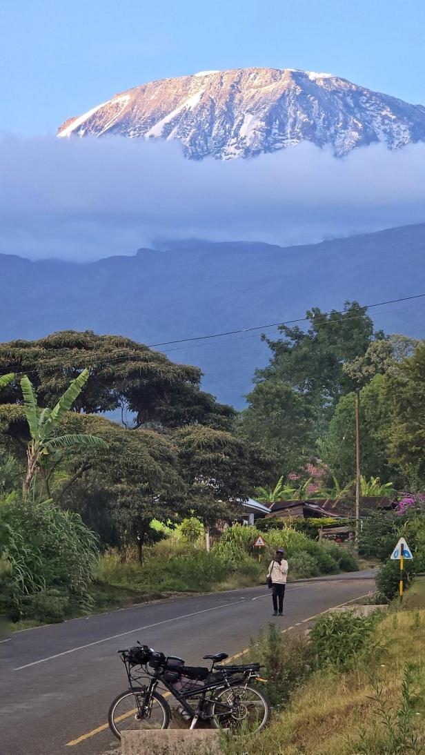 Mann mit Fahrrad und drei Kindern am weißen Sandstrand vor türkisblauem Meer und Segelbooten.