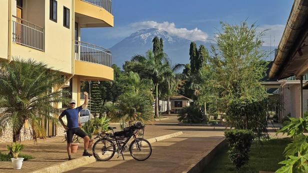 Mann mit Fahrrad und drei Kindern am weißen Sandstrand vor türkisblauem Meer und Segelbooten.