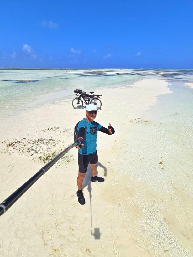 Mann mit Fahrrad und drei Kindern am weißen Sandstrand vor türkisblauem Meer und Segelbooten.