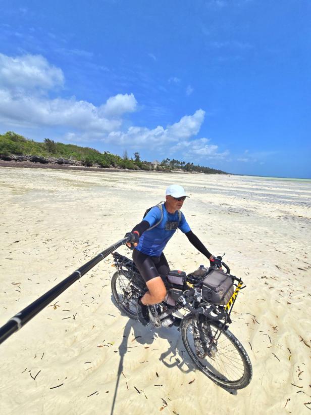 Mann mit Fahrrad und drei Kindern am weißen Sandstrand vor türkisblauem Meer und Segelbooten.
