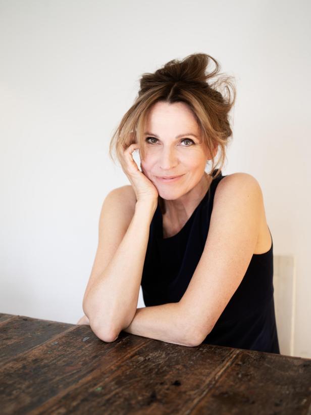 Frau mit locker hochgesteckten Haaren sitzt lächelnd an einem Holztisch und stützt den Kopf auf die Hand.