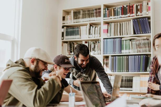 Mehrere junge Menschen sitzen lachend an einem Tisch mit Laptops in einer Bibliothek mit Bücherregalen im Hintergrund.