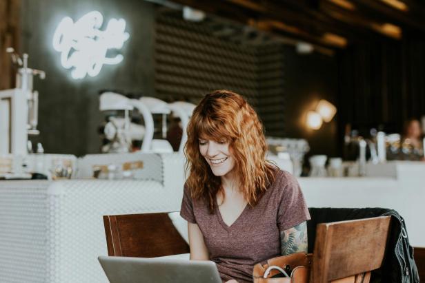 Eine junge Frau mit roten Haaren sitzt lächelnd an einem Tisch in einem Café und arbeitet an einem Laptop.