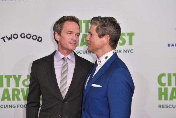 Neil Patrick Harris und David Burtka stehen sich vor einem City Harvest-Hintergrund gegenüber und schauen sich an.