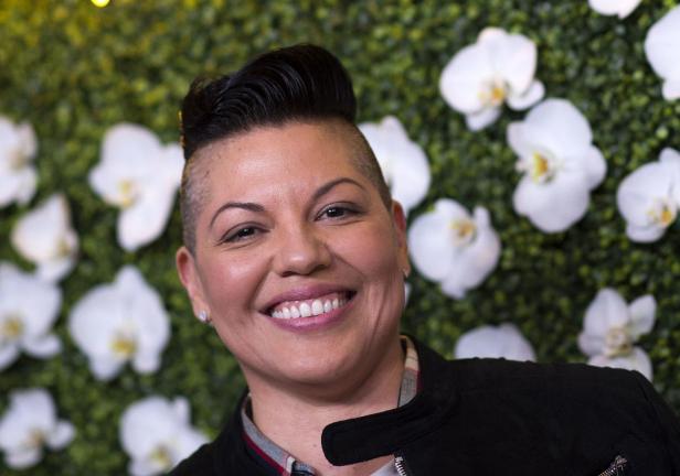 Sara Ramirez lächelt vor einer Wand aus grünem Blattwerk und weißen Orchideen.