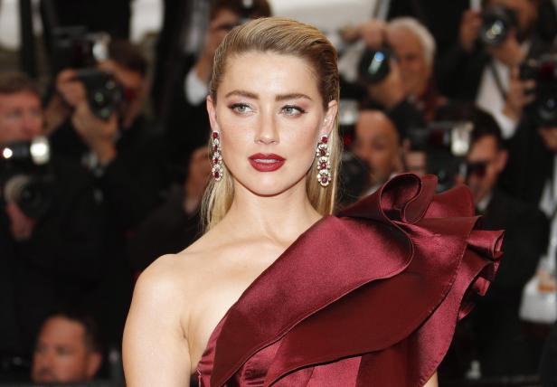 Amber Heard trägt ein dunkelrotes Kleid mit Rüschen und auffällige Ohrringe vor einer Kulisse aus Fotografen.