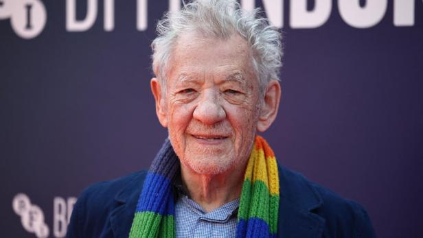 Ian McKellen lächelt vor einem dunklen Hintergrund und trägt einen Schal in Regenbogenfarben.