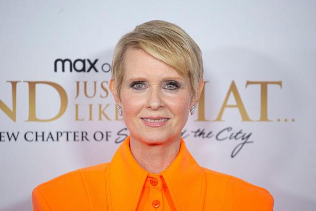 Cynthia Nixon lächelt vor einem Hintergrund mit dem Logo der Serie „And Just Like That...“ und trägt ein leuchtend orangefarbenes Oberteil.