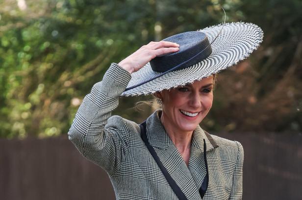Prinzessin Kate hält ihren Hut mit breiter Krempe und lächelt in einem karierten Blazer.