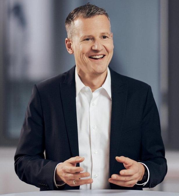 Ein Mann im Anzug und weißem Hemd lächelt und gestikuliert mit den Händen in einem modernen Innenraum.