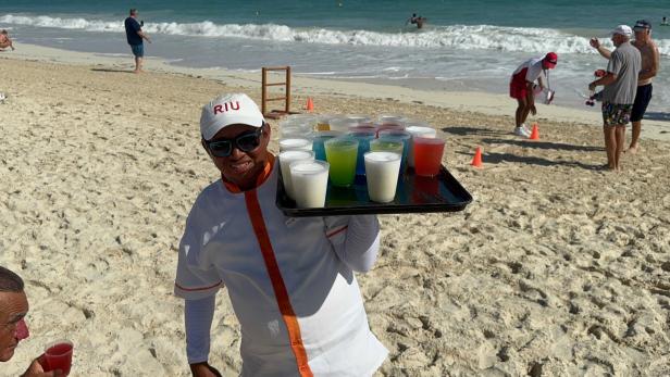 Ein Mann mit Sonnenbrille und Tablett voller bunter Getränke steht am Sandstrand, im Hintergrund spielen Menschen am Meer.