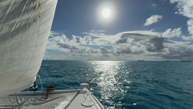 Ein Segelboot fährt auf dem glitzernden, blauen Meer unter einem bewölkten Himmel.