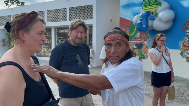 Vier Personen stehen im Freien, ein Mann mit Stirnband berührt eine Frau an der Schulter, im Hintergrund bunte Wandmalerei.