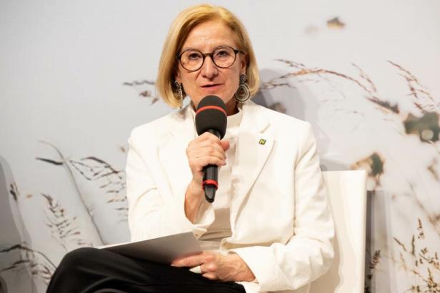 Frau im weißen Blazer spricht mit Mikrofon vor Hintergrund mit Gräsern.