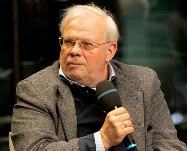Ein älterer Mann mit Brille hält ein Mikrofon und spricht bei einer Podiumsdiskussion.