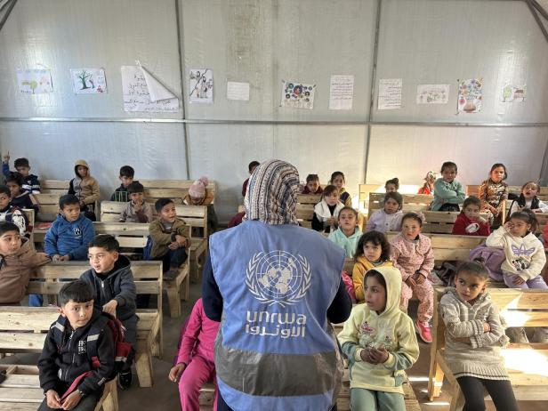 Eine Frau mit UNRWA-Weste spricht vor einer Gruppe von Kindern, die auf Holzbänken in einem einfachen Klassenraum sitzen.