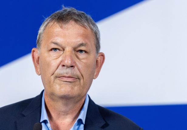 Ein Mann mit grauen Haaren und Anzug steht vor einem blau-weiß gemusterten Hintergrund.