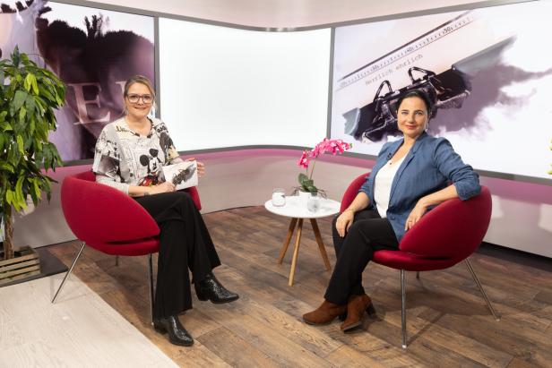 Zwei Frauen sitzen in roten Sesseln in einem modernen Studio, zwischen ihnen ein kleiner Tisch mit einer Orchidee und zwei Gläsern Wasser.