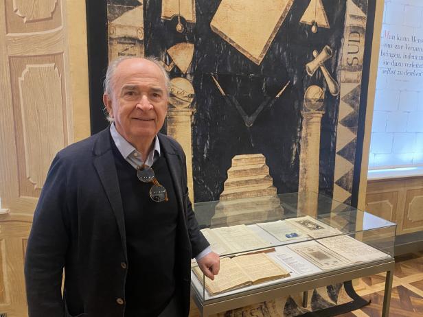 Ein Mann steht vor einer Vitrine mit Dokumenten und einem Wandteppich mit freimaurerischen Symbolen.
