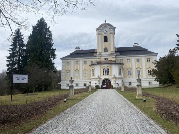 Ein gelbes Schloss mit Uhrturm und gepflastertem Weg, umgeben von Bäumen und einem Schild zum Freimaurer-Museum.