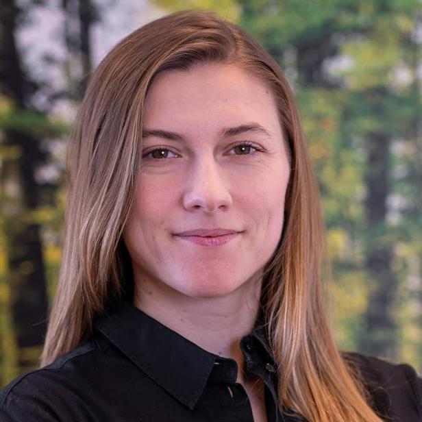 Junge Frau mit langen, glatten, blonden Haaren und schwarzem Hemd steht vor unscharfem Waldhintergrund.