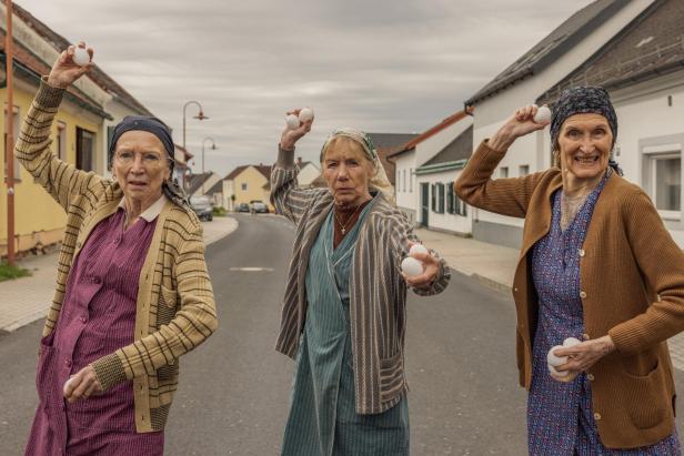 Drei ältere Frauen stehen auf einer Straße und halten Eier in Wurfposition, mit entschlossenen Gesichtsausdrücken.