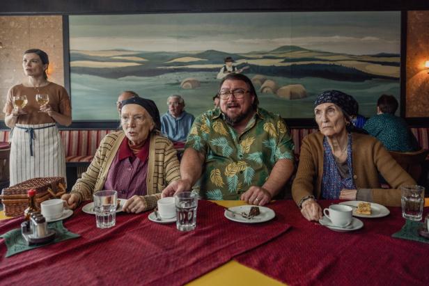 Drei Personen sitzen an einem Tisch mit Kuchen und Getränken, im Hintergrund serviert eine Frau zwei Gläser Weißwein.