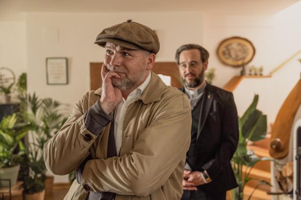 Ein nachdenklicher Mann mit Mütze steht im Vordergrund, im Hintergrund steht ein Mann im Anzug mit Priesterkragen.