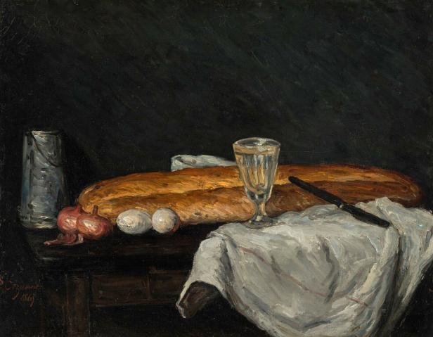 Ein langes Brot, zwei Zwiebeln, zwei Eier, ein Glas Wasser und ein Messer liegen auf einem Tuch auf einem dunklen Tisch.