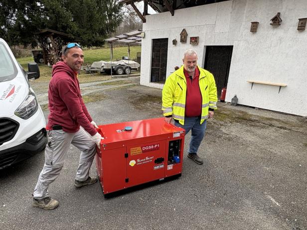 Zwei Männer bewegen gemeinsam einen roten Dieselgenerator auf einem Hof vor einem Gebäude mit Vogelhäusern an der Wand.
