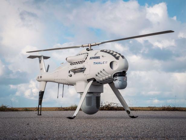 Eine weiße unbemannte Hubschrauberdrohne mit Kamerasensoren steht auf Asphalt vor bewölktem Himmel.