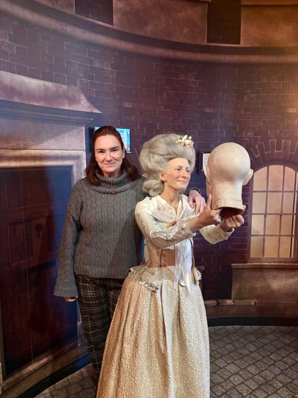 Eine Frau posiert neben einer Wachsfigur im historischen Kleid, die einen Kopf hält.