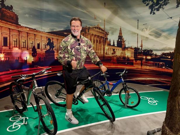 Arnold Schwarzenegger posiert auf einem Fahrradweg vor einem Wien-Hintergrund und zeigt den Daumen nach oben.