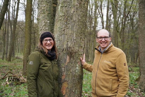 Katharina Leb und Lukas Wurzinger vom Unternehmen Klosterwald stehen lächelnd an einem Baum im Bestattungswald.