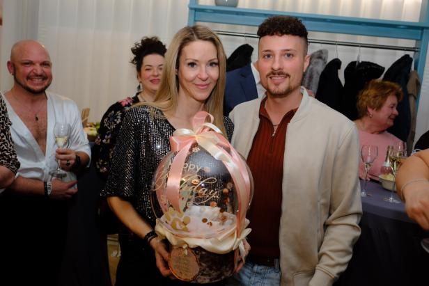 Eine Frau und ein Mann halten gemeinsam ein großes, dekoriertes Geburtstagsgeschenk, umgeben von feiernden Menschen.