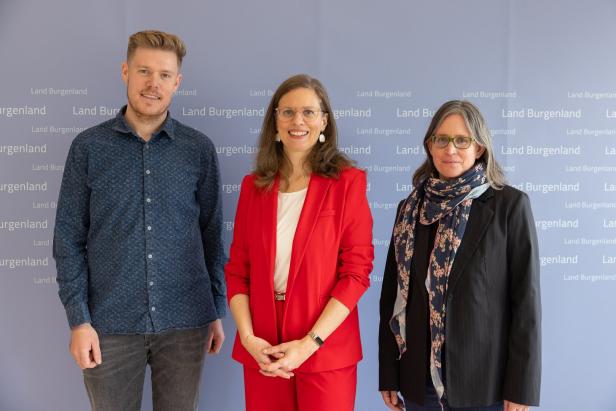 Drei Personen stehen nebeneinander vor einer hellblauen Wand mit der Aufschrift „Land Burgenland“.