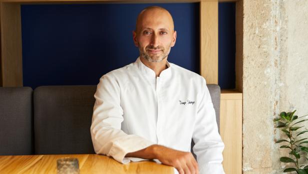 Ein Koch im weißen Jacket sitzt an einem Holztisch vor einer blauen Wand.