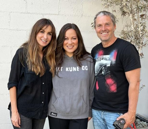 Drei lächelnde Personen posieren vor einer hellen Wand, eine Frau trägt einen grauen Hoodie.