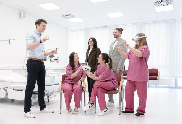 Mehrere Personen in einem hellen Raum, einige in pinker Kleidung, testen Virtual-Reality-Brillen, während andere zuschauen und erklären.