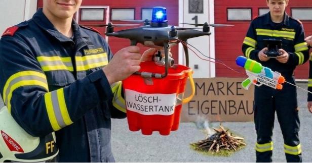 Zwei Feuerwehrleute mit einer Drohne, an der ein Wassereimer und eine Wasserpistole befestigt sind, vor einem kleinen Lagerfeuer.