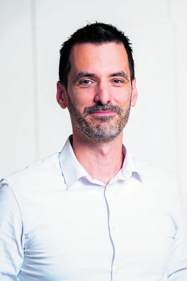 Mann mit dunklen Haaren und Bart trägt ein weißes Hemd und lächelt leicht vor hellem Hintergrund.