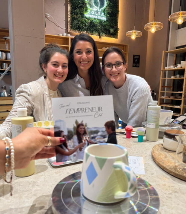 Drei Frauen sitzen an einem Tisch und lächeln.