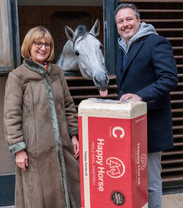 Zwei Personen stehen vor dem Fenster einer Stallung, ein Pferd reckt den Kopf aus der Öffnung und streckt die Zunge nach einem Sack mit Holzflocken aus.