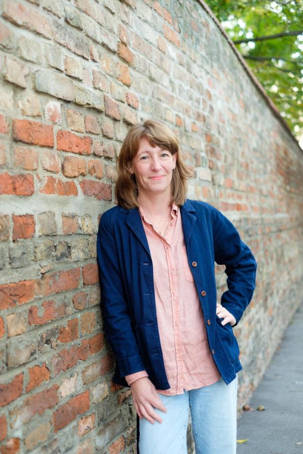 Barbara Nothegger lehnt entspannt an einer Backsteinmauer und blickt in die Kamera.